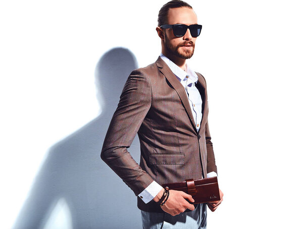 portrait of handsome fashion stylish hipster businessman model dressed in elegant brown suit in sunglasses posing near white wall in studio. Pulls out or put his leather wallet into jacket pocket