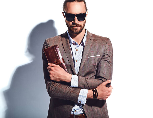 portrait of handsome fashion stylish hipster businessman model dressed in elegant brown suit in sunglasses posing near white wall in studio. Pulls out or put his leather wallet into jacket pocket