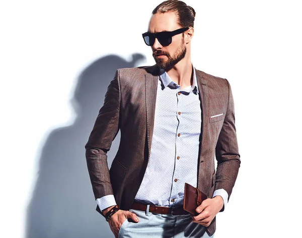yakışıklı şık hipster işadamı manken portresi Studio beyaz duvarının yakınında poz güneş gözlüklü zarif kahverengi takım elbise giymiş. Çıkarır veya onun deri cüzdan ceket cebine attı