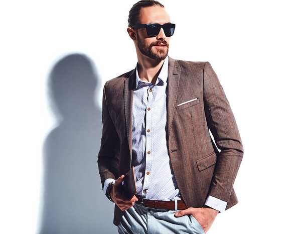 portrait of handsome fashion stylish hipster businessman model dressed in elegant brown suit in sunglasses posing near white wall in studio. Pulls out or put his leather wallet into jacket pocket
