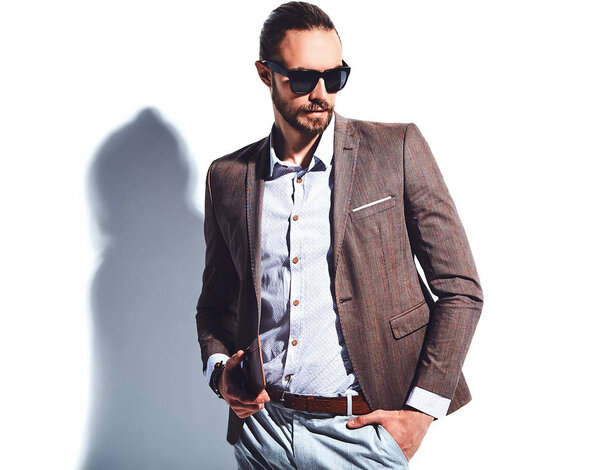 portrait of handsome fashion stylish hipster businessman model dressed in elegant brown suit in sunglasses posing near white wall in studio. Pulls out or put his leather wallet into jacket pocket