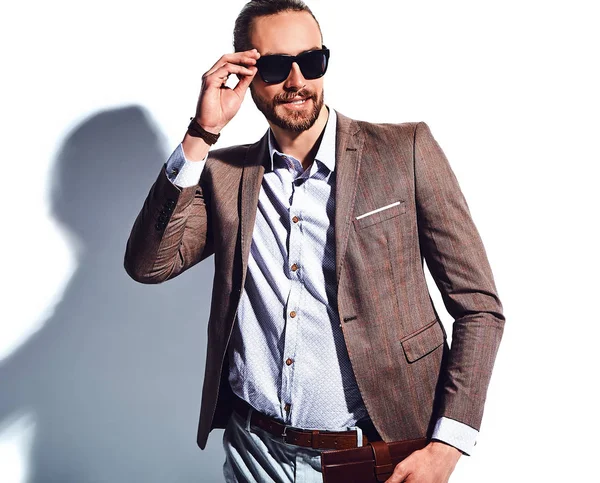 yakışıklı şık hipster işadamı manken portresi Studio beyaz duvarının yakınında poz güneş gözlüklü zarif kahverengi takım elbise giymiş. Çıkarır veya onun deri cüzdan ceket cebine attı