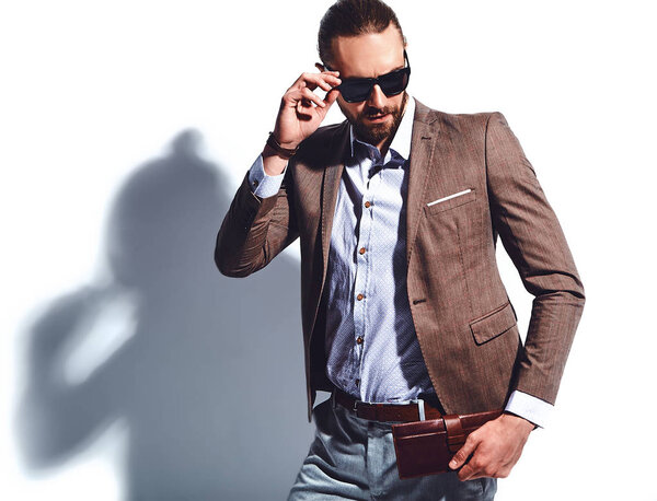 portrait of handsome fashion stylish hipster businessman model dressed in elegant brown suit in sunglasses posing near white wall in studio. Pulls out or put his leather wallet into jacket pocket