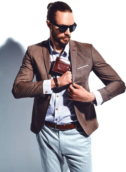 Portrait of handsome fashion stylish hipster businessman model dressed in elegant brown suit in sunglasses posing near white wall in studio. Pulls out or put his leather wallet into jacket pocket — Stock Photo, Image