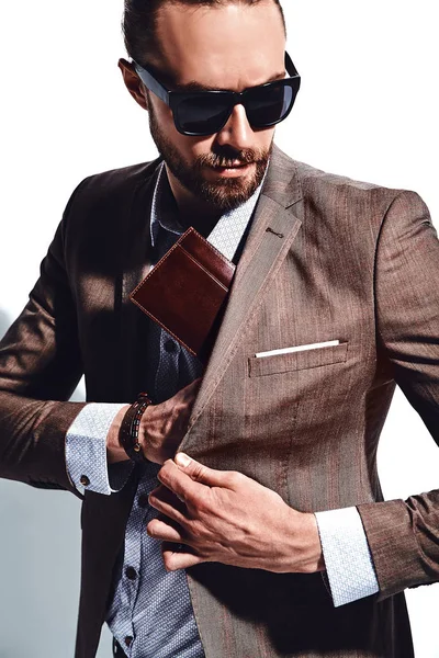 portrait of handsome fashion stylish hipster businessman model dressed in elegant brown suit in sunglasses posing near white wall in studio. Pulls out or put his leather wallet into jacket pocket