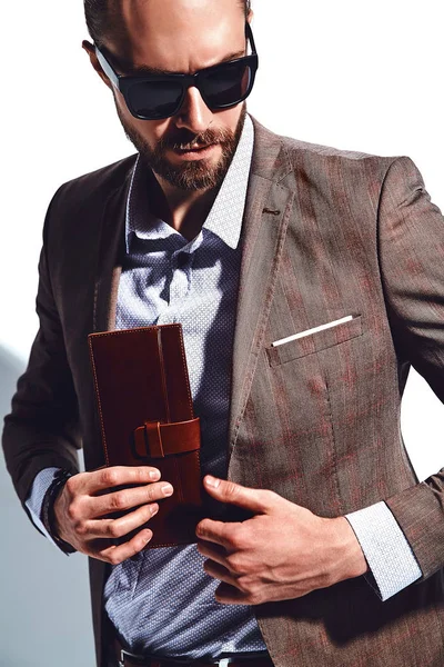 portrait of handsome fashion stylish hipster businessman model dressed in elegant brown suit in sunglasses posing near white wall in studio. Pulls out or put his leather wallet into jacket pocket