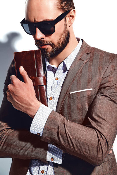 portrait of handsome fashion stylish hipster businessman model dressed in elegant brown suit in sunglasses posing near white wall in studio. Pulls out or put his leather wallet into jacket pocket