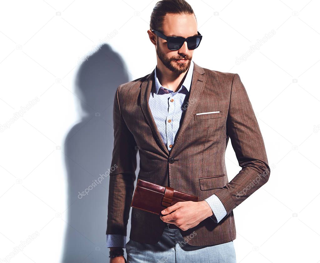 portrait of handsome fashion stylish hipster businessman model dressed in elegant brown suit in sunglasses posing near white wall in studio. Pulls out or put his leather wallet into jacket pocket