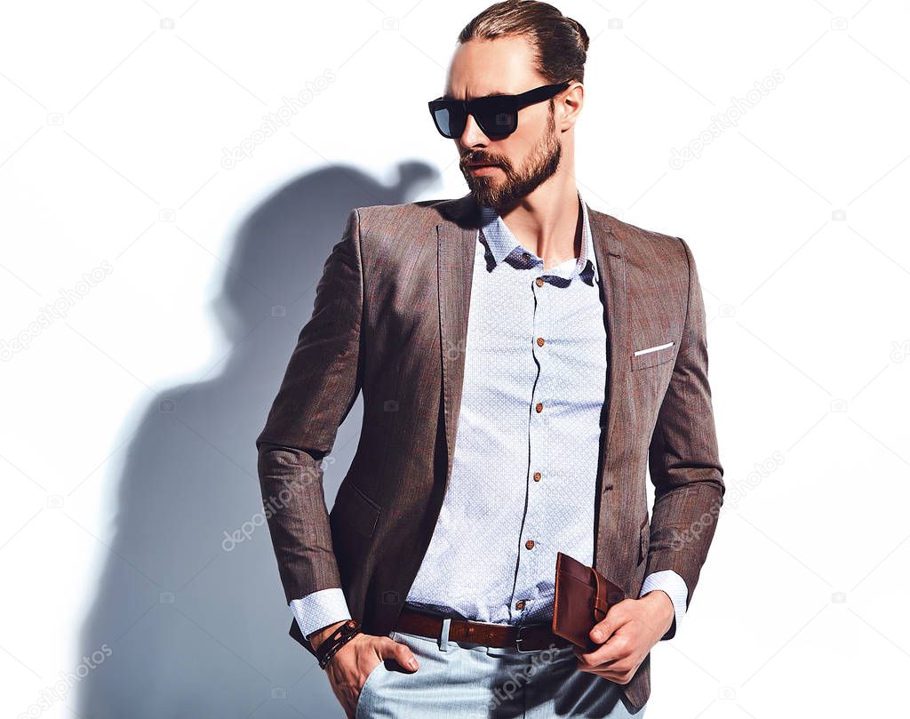 portrait of handsome fashion stylish hipster businessman model dressed in elegant brown suit in sunglasses posing near white wall in studio. Pulls out or put his leather wallet into jacket pocket