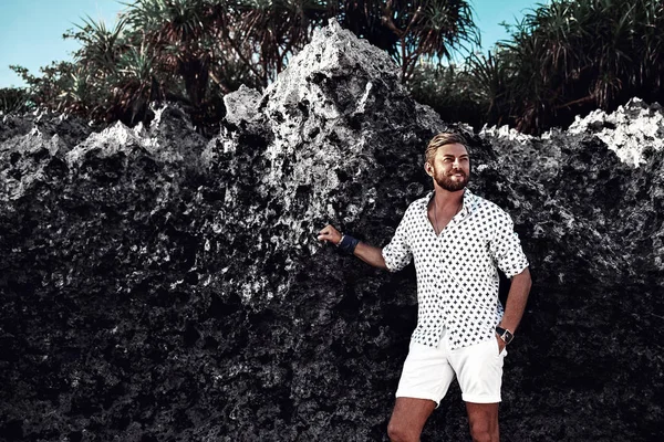 Ritratto di un bel modello di uomo di moda che indossa vestiti bianchi in posa vicino a rocce sulla spiaggia — Foto Stock