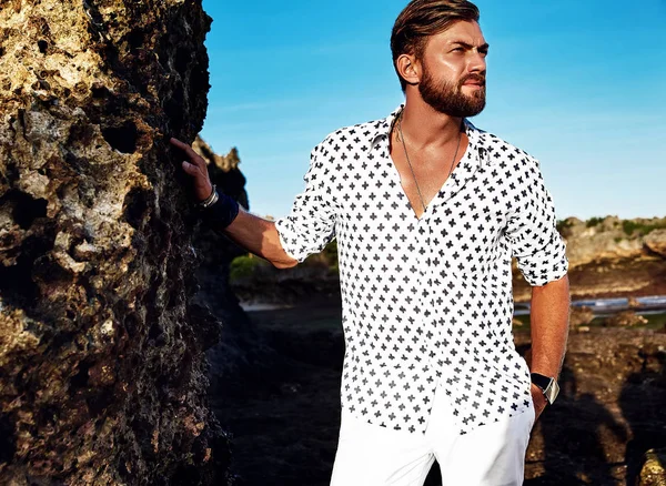 Retrato del guapo modelo de hombre de moda con ropa blanca posando cerca de las rocas en la playa —  Fotos de Stock