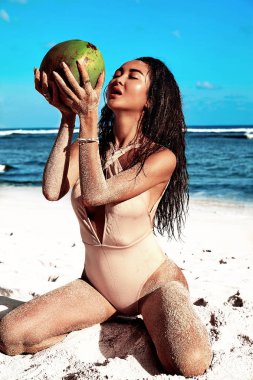 güzel beyaz sunbathed kadın portresi modeli bej mayo mavi gökyüzü ve okyanus zemin üzerine beyaz kum ile yaz sahilde poz karanlık uzun saçlı. Taze hindistan cevizi içme