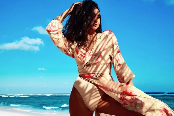 Retrato de hermosa mujer caucásica bronceada modelo con pelo largo oscuro en vestido volador beige posando en la playa de verano con arena blanca sobre el cielo azul y el fondo del océano. Tocando su cabello —  Fotos de Stock