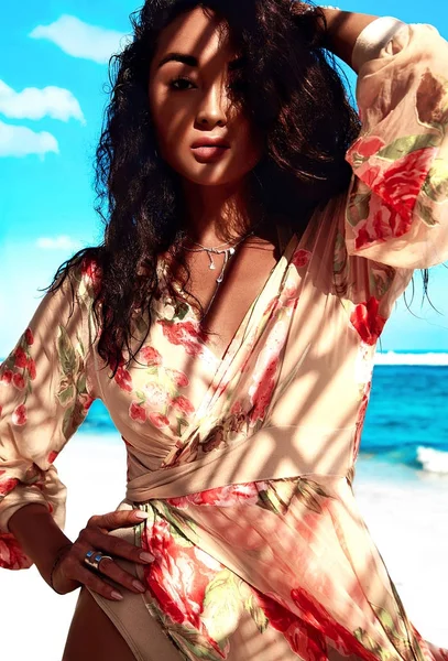 Portrait of beautiful caucasian sunbathed woman model with dark long hair in beige flying dress posing on summer beach with white sand on blue sky and ocean background. Touching her hair — Stock Photo, Image