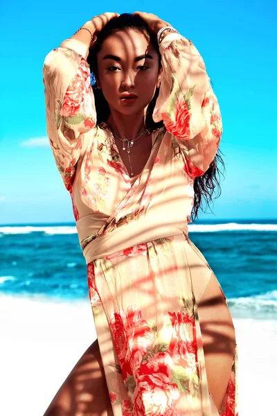 Retrato de hermosa mujer caucásica bronceada modelo con pelo largo oscuro en vestido volador beige posando en la playa de verano con arena blanca sobre el cielo azul y el fondo del océano. Tocando su cabello —  Fotos de Stock