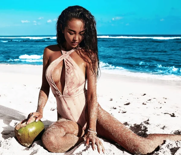 Porträtt av vackra kaukasiska solsvedda kvinna modell med mörkt långt hår i beige baddräkt poserar på sommar strand med vit sand på blå himmel och hav bakgrund. Dricka färsk kokosnöt — Stockfoto