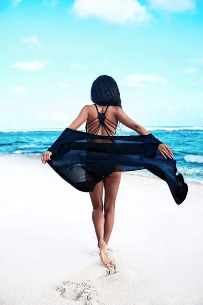 Portrait of beautiful caucasian sunbathed woman model with dark long hair in black  swimsuit with flying cape posing on summer beach with white sand on blue sky and ocean background — Stock Photo, Image