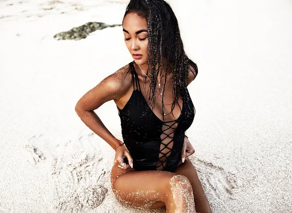 Porträtt av vackra kaukasiska solsvedda kvinna modell med mörkt långt hår i svart baddräkt poserar på sommar strand med vit sand på blå himmel och hav bakgrund — Stockfoto