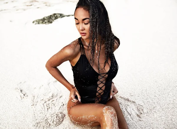 Portrait of beautiful caucasian sunbathed woman model with dark long hair in black  swimsuit  posing on summer beach with white sand on blue sky and ocean background — Stock Photo, Image