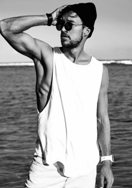 Adam manken beyaz T-shirt ve mavi okyanus ve gökyüzü arka plan üzerinde poz güneş gözlüğü rahat kıyafetler giymiş yakışıklı hipster portresi sunbathed