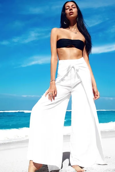 Retrato da bela mulher branca modelo com cabelos longos escuros em calças clássicas de perna larga posando na praia de verão com areia branca no céu azul e fundo do oceano — Fotografia de Stock