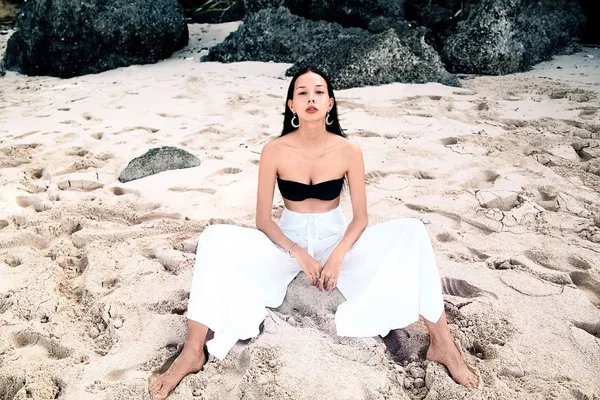 Portrait of beautiful caucasian woman model with dark long hair in wide-leg classic pants  sittting on summer beach with white sand near rocks — Stock Photo, Image