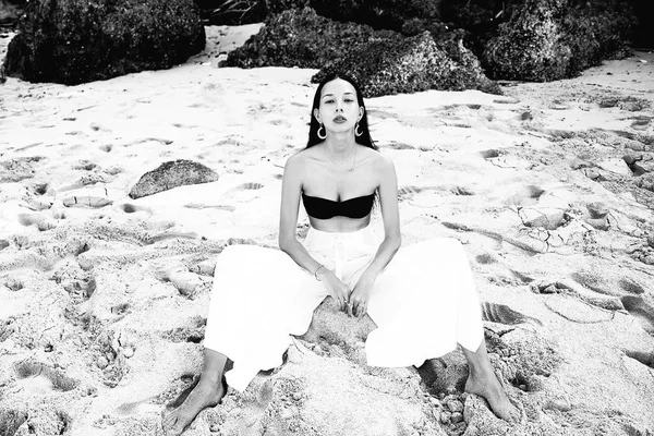 Retrato da bela mulher branca modelo com cabelos longos escuros em calças clássicas de perna larga sentado na praia de verão com areia branca perto de rochas — Fotografia de Stock