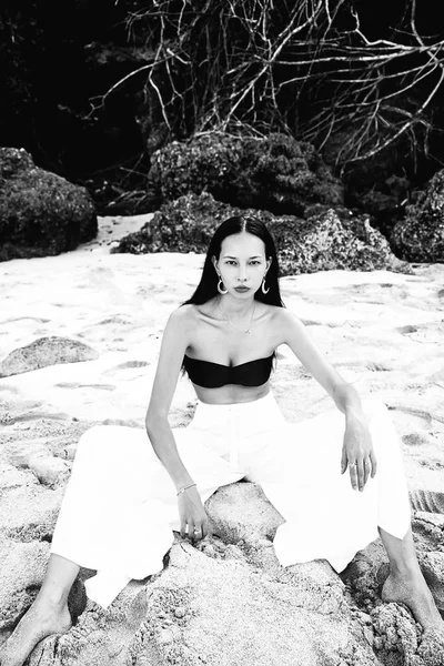 Πορτρέτο του όμορφη γυναίκα Καυκάσιος μοντέλο με σκούρα μακριά μαλλιά σε κλασικό παντελόνι καμπάνα sittting καλοκαίρι παραλία με λευκή άμμο κοντά σε βράχους — Φωτογραφία Αρχείου