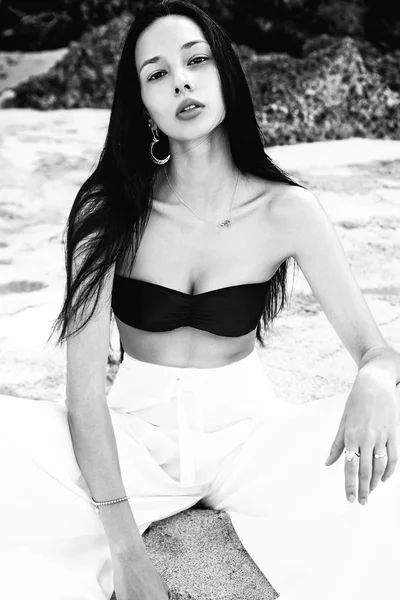 Portrait of beautiful caucasian woman model with dark long hair in wide-leg classic pants  sittting on summer beach with white sand near rocks — Stock Photo, Image