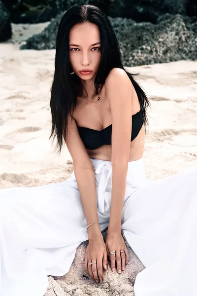 Portrait of beautiful caucasian woman model with dark long hair in wide-leg classic pants  sittting on summer beach with white sand near rocks — Stock Photo, Image