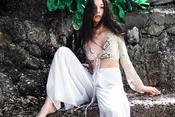 Portrait of beautiful caucasian woman model with dark long hair in wide-leg classic pants  posing near rocks and green tropical exotic leaves background — Stock Photo, Image