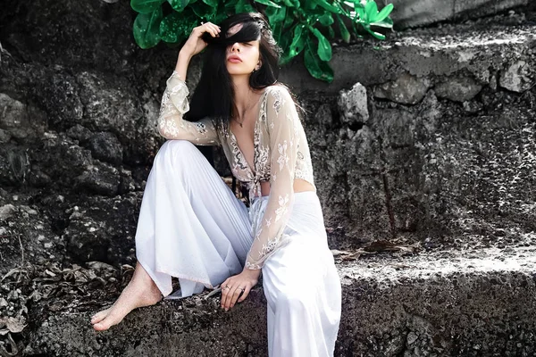 Portrait of beautiful caucasian woman model with dark long hair in wide-leg classic pants  posing near rocks and green tropical exotic leaves background — Stock Photo, Image