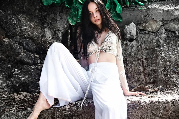 Retrato de bela mulher branca modelo com cabelos longos escuros em calças clássicas de perna larga posando perto de rochas e verde tropical exótico folhas fundo — Fotografia de Stock