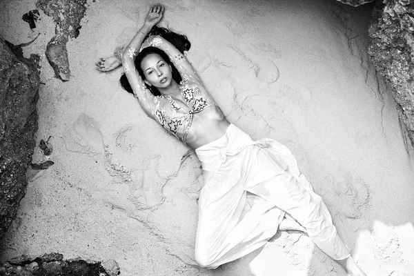 Retrato de belo modelo de mulher caucasiana com cabelos longos escuros em calças clássicas de perna larga deitado na areia branca na praia perto de rochas. Vista superior — Fotografia de Stock