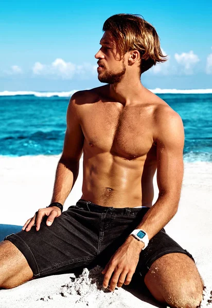 Retrato del guapo modelo de hombre de moda bañado por el sol posando en la playa de verano con arena blanca sobre el cielo azul y el fondo del océano — Foto de Stock