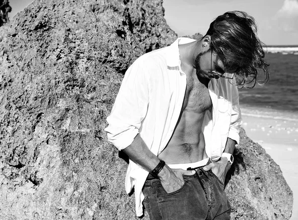 Portrait of handsome sunbathed fashion man model wearing white shirt clothes in glasses posing near rocks on summer beach on blue sky and ocean background — Stock Photo, Image