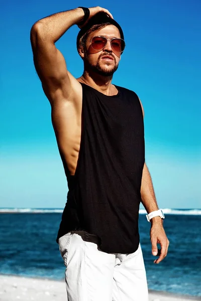 Ritratto di bello hipster abbronzato modello uomo di moda indossando abiti casual in nero T-shirt e occhiali da sole in posa su cielo blu e sfondo oceano. Toccandogli la testa — Foto Stock