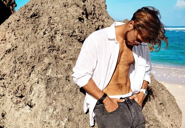 ハンサムな日光浴ファッション男のポートレート モデル グラスの青い空と海の背景に夏のビーチの岩の近くのポーズで服を着て白いシャツ — ストック写真