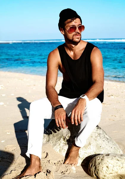 Portrait of handsome hipster sunbathed fashion man model wearing casual clothes in black T-shirt and sunglasses sitting on rocks — Stock Photo, Image