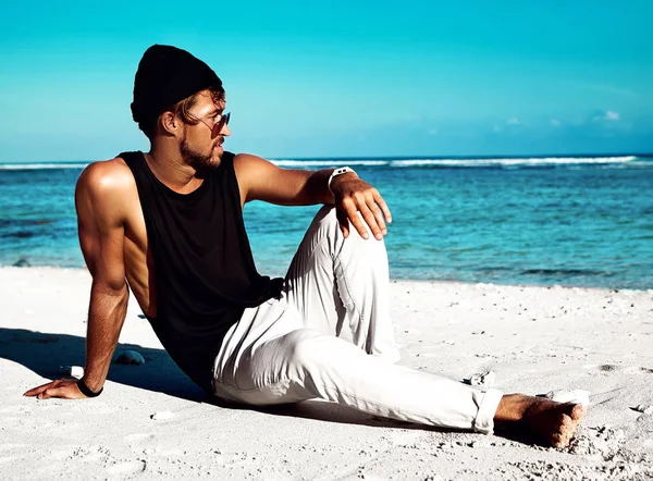 Portrait of handsome hipster sunbathed fashion man model wearing casual clothes in black T-shirt and sunglasses sitting on white sand near blue ocean and sky background — Stock Photo, Image