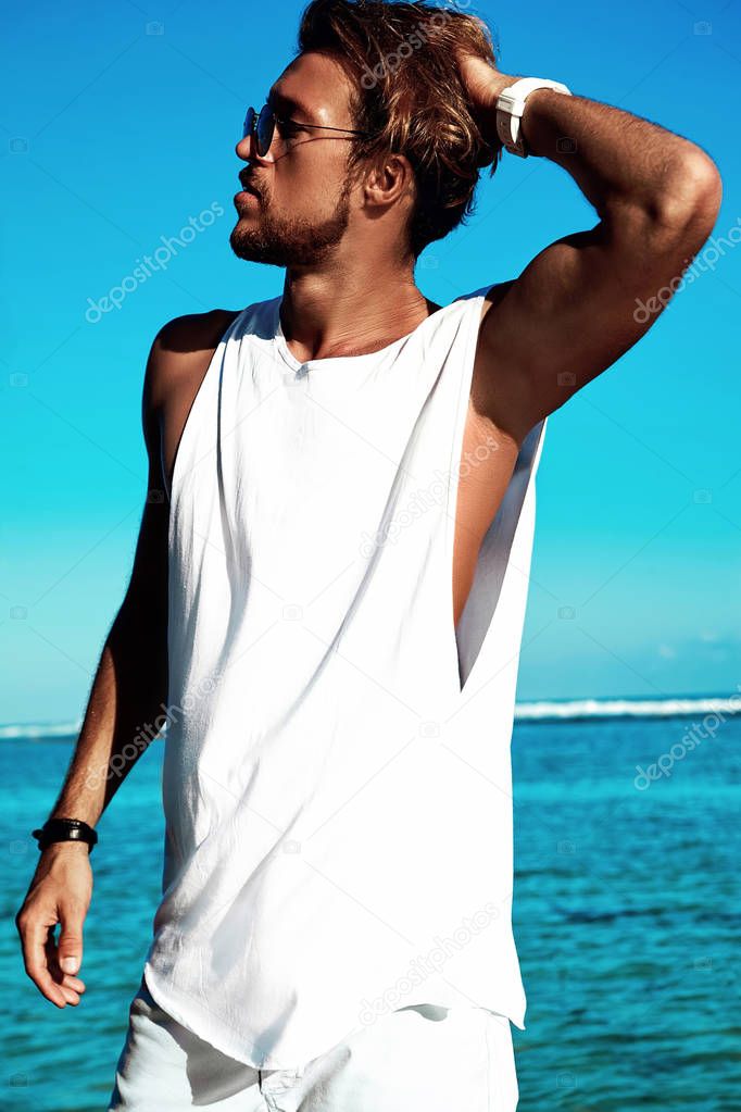 Portrait of handsome hipster sunbathed fashion man model wearing casual clothes in white T-shirt and sunglasses posing on blue ocean and sky background