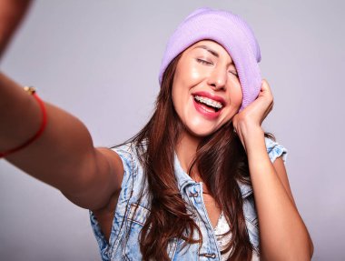 Güzel gülümseyen şirin esmer kadın modeli rahat yaz kot giysileri mor beanie smartphone üzerinde gri izole selfie fotoğraf yapma hiç makyaj ile portresi