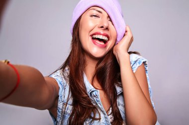 Güzel gülümseyen şirin esmer kadın modeli rahat yaz kot giysileri mor beanie smartphone üzerinde gri izole selfie fotoğraf yapma hiç makyaj ile portresi