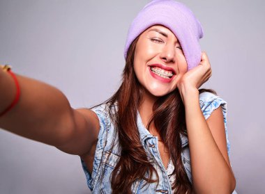 Güzel gülümseyen şirin esmer kadın modeli rahat yaz kot giysileri mor beanie smartphone üzerinde gri izole selfie fotoğraf yapma hiç makyaj ile portresi