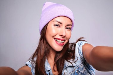 Güzel gülümseyen şirin esmer kadın modeli rahat yaz kot giysileri mor beanie smartphone üzerinde gri izole selfie fotoğraf yapma hiç makyaj ile portresi