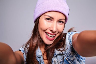 Güzel gülümseyen şirin esmer kadın modeli rahat yaz kot giysileri mor beanie smartphone üzerinde gri izole selfie fotoğraf yapma hiç makyaj ile portresi
