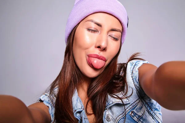Mor beanie smartphone üzerinde gri izole selfie fotoğraf yapma hiç makyaj ile rahat yaz kot giysileri güzel şirin esmer kadın modeli portresi. Dilini gösterilen