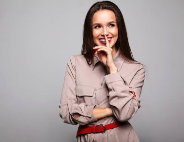 Güzel gülümseyen şirin esmer kadın model parlak makyaj ve gri izole kırmızı dudaklar ile rahat bej şık kıyafetler içinde portresi. Dudaklarında parmak yerleştirme 