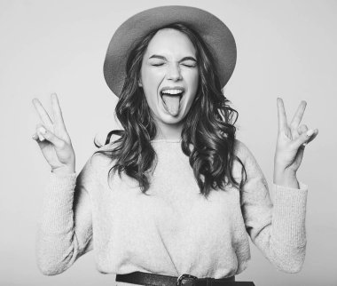 Genç şık gülen kız modeli gri rahat yaz giysileri ile doğal makyaj gri arka plan üzerinde izole kahverengi şapkalı portresi. Kameraya bakarak ve barış işareti ve dilini gösteren