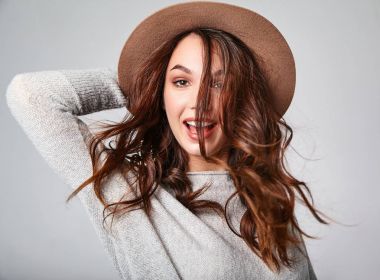 Genç şık gülen kız modeli gri rahat yaz giysileri doğal makyaj ve uzun saç gri arka plan üzerinde izole üfleme ile kahverengi şapkalı portresi. Kameraya bakarak